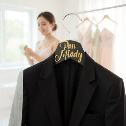 An elegant, modern wedding preparation scene in a brightly lit bridal suite, featuring a wooden hanger engraved with 'Pan Młody' in gold lettering The hanger holds a crisp, impeccably tailored black tuxedo j
